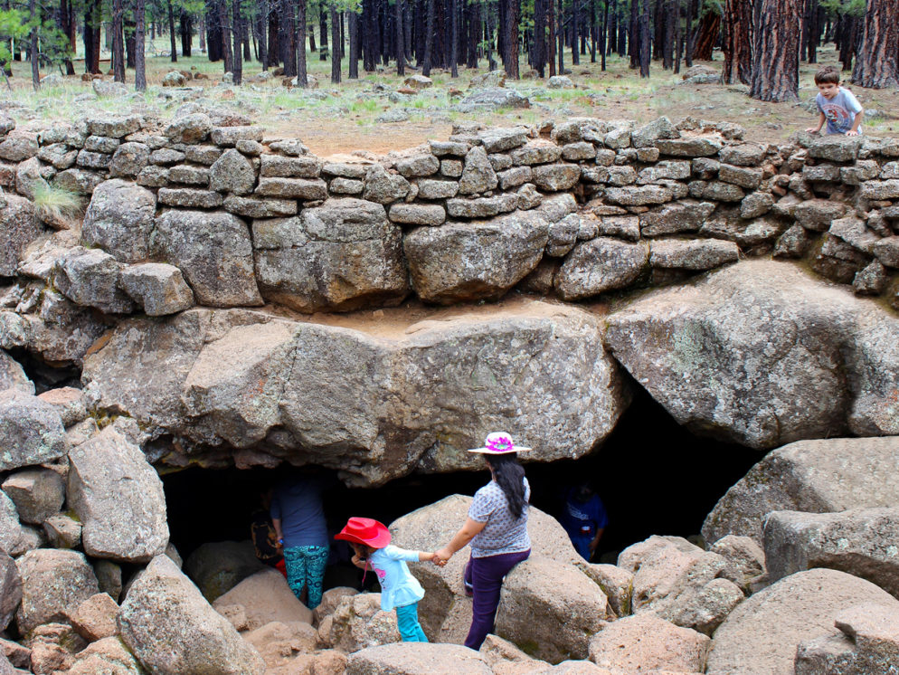 A family standing on the boulders outside the opening of the Lava River Tube, on the Lava River Tube Trail, near Flagstaff, Arizona. Easy Arizona hiking trails. Flagstaff area hiking trails. Arizona family friendly hiking trails. Copyright azutopia. No use without permission.