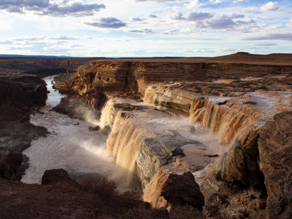 View of water rushing over the top of Grand Falls; With the canyon and Little Colorado River below; Sunset; Shafts of Light; Chocolate Falls; Leupp; Navajo Nation; Painted Desert; Arizona; Spring Runoff; Blue Sky; Moderate Hiking Trails; Northern Arizona; Family Friendly Hiking Trails; Copyright azutopia.com; No use without permission.
