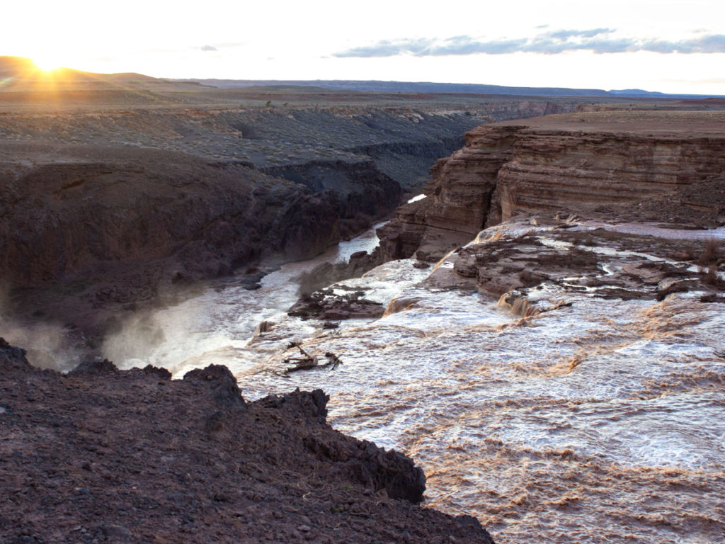 View of water rushing over the top of Grand Falls; With the canyon and Little Colorado River below; Sunset; Chocolate Falls; Leupp; Navajo Nation; Painted Desert; Arizona; Spring Runoff; Blue Sky; Moderate Hiking Trails; Northern Arizona; Family Friendly Hiking Trails; Copyright azutopia.com; No use without permission.