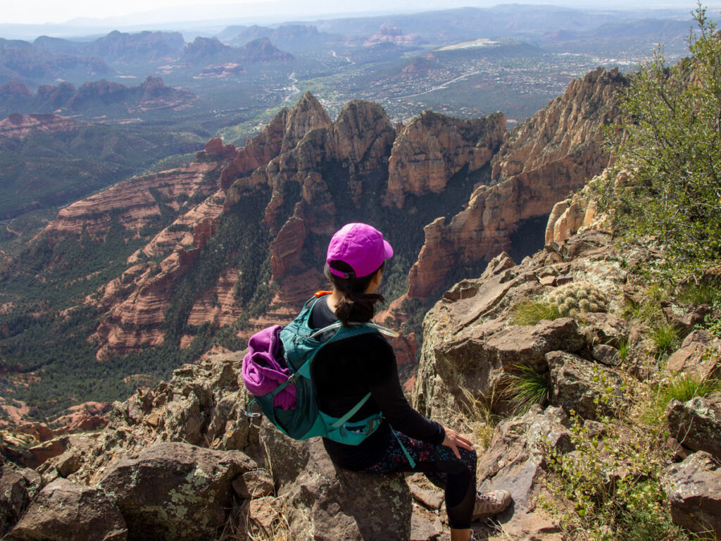 Hiker sitting on the edge, at the top of Wilson Mountain, before a westward view over Sedona, Arizona's Red Rock Cliffs, Oak Creek Canyon, the Sedona Airport and the city. Taken from the Wilson Mountain Hiking Trail. Difficult Hiking Trails. Dog Friendly Hiking Trails. Copyright azutopia.com. No use without permission.